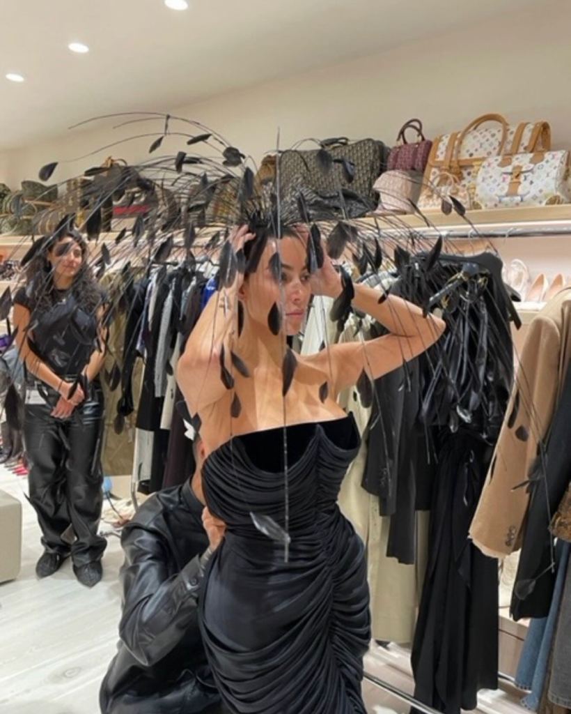 Kim Kardashian wearing a black strapless dress and a large feathery headpiece, being attended to by someone adjusting her dress, with another person in black leather behind her, and shelves of handbags in the background.