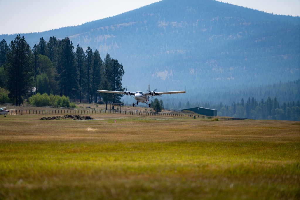 A Twin Otter airplane takes off over a field with a pine tree forest and a mountain in the background.