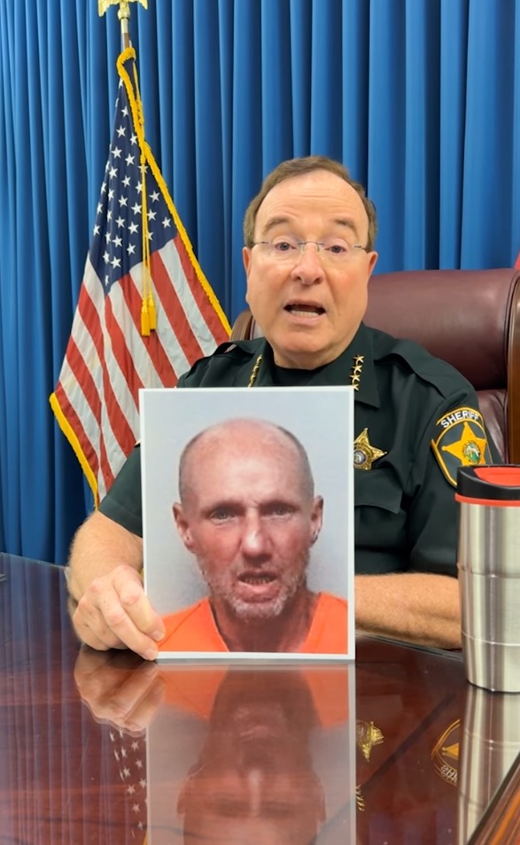 A police officer in glasses, wearing a green uniform with badges, holds up a mugshot of a bald man in an orange jumpsuit.