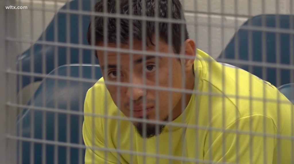 Rosali Fernandez-Cruz, a man with dark hair and a short beard, wearing a yellow shirt, looks out from behind a metal grate.