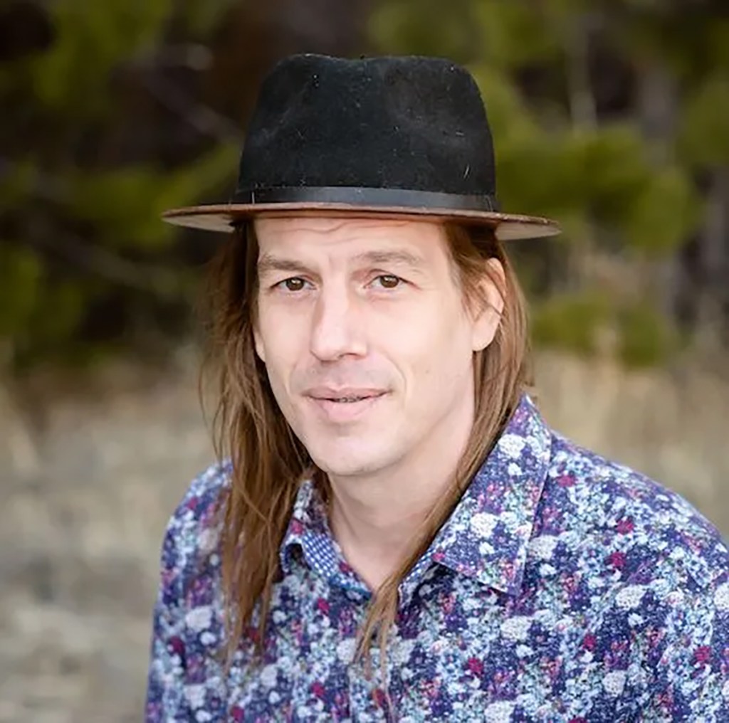 A man with long brown hair, wearing a black hat and a floral patterned shirt.