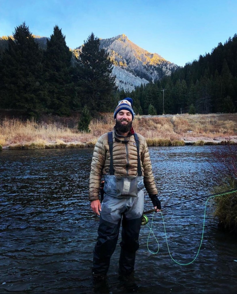 Charlie Eppard in fishing waders in a river with a mountain and forest in the background.