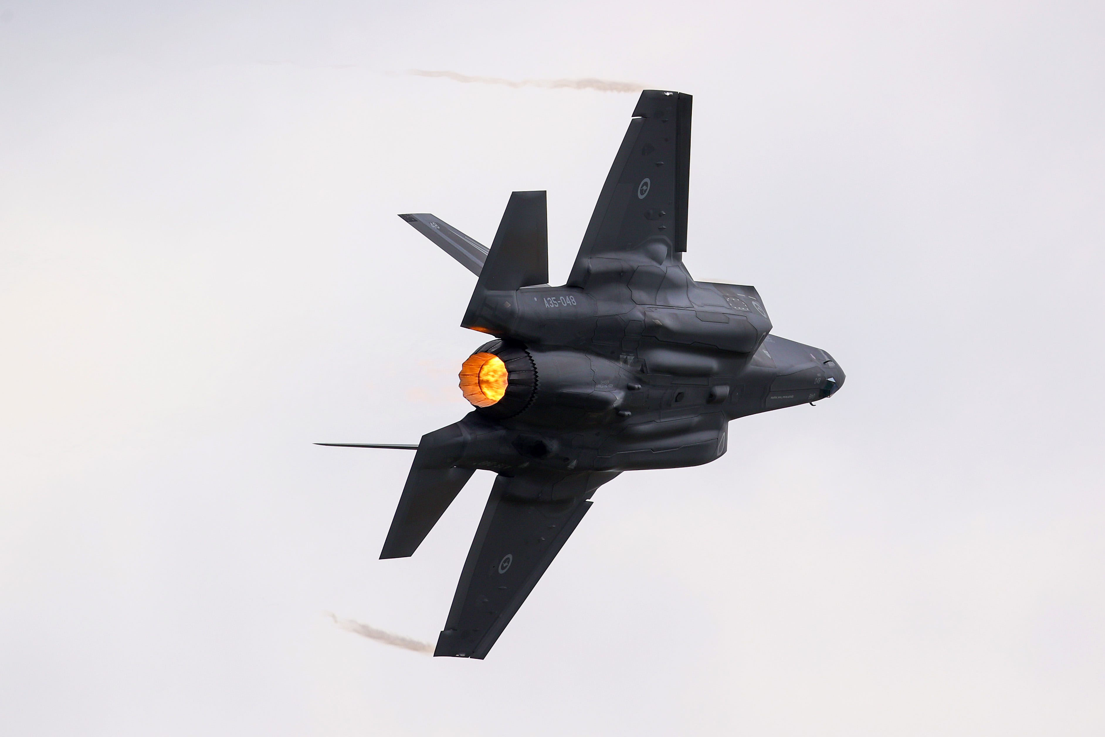 A grey jet on its side in the sky with orange flame visable in its tail