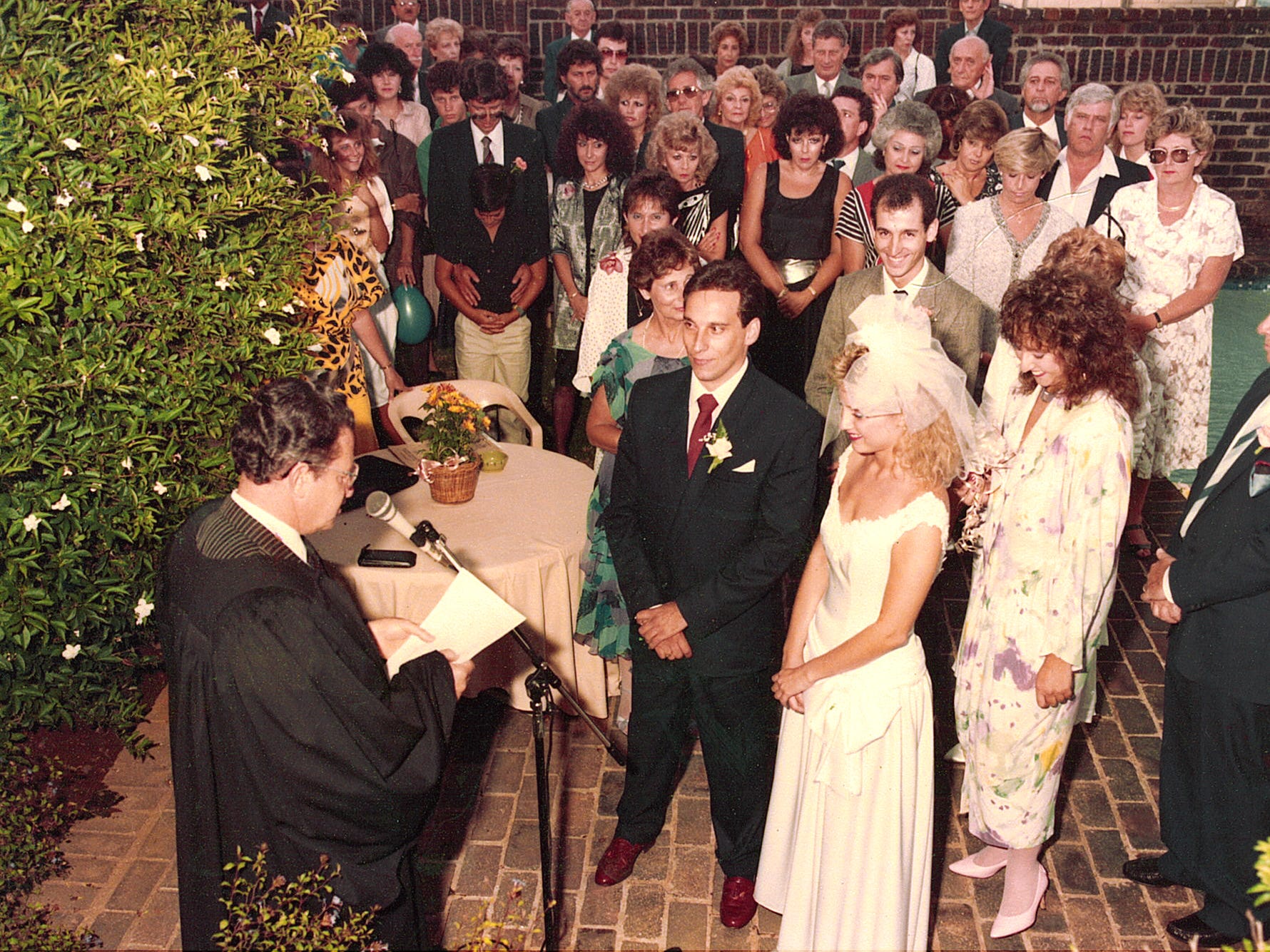 Couple in wedding attire standing in front of man in robes with microphone and guests crowded behind them
