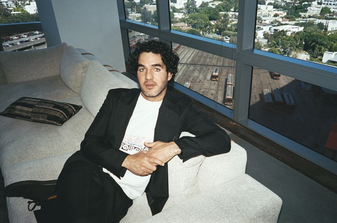 A color photograph of Benny Safdie in a white tee and black blazer seated on a couch