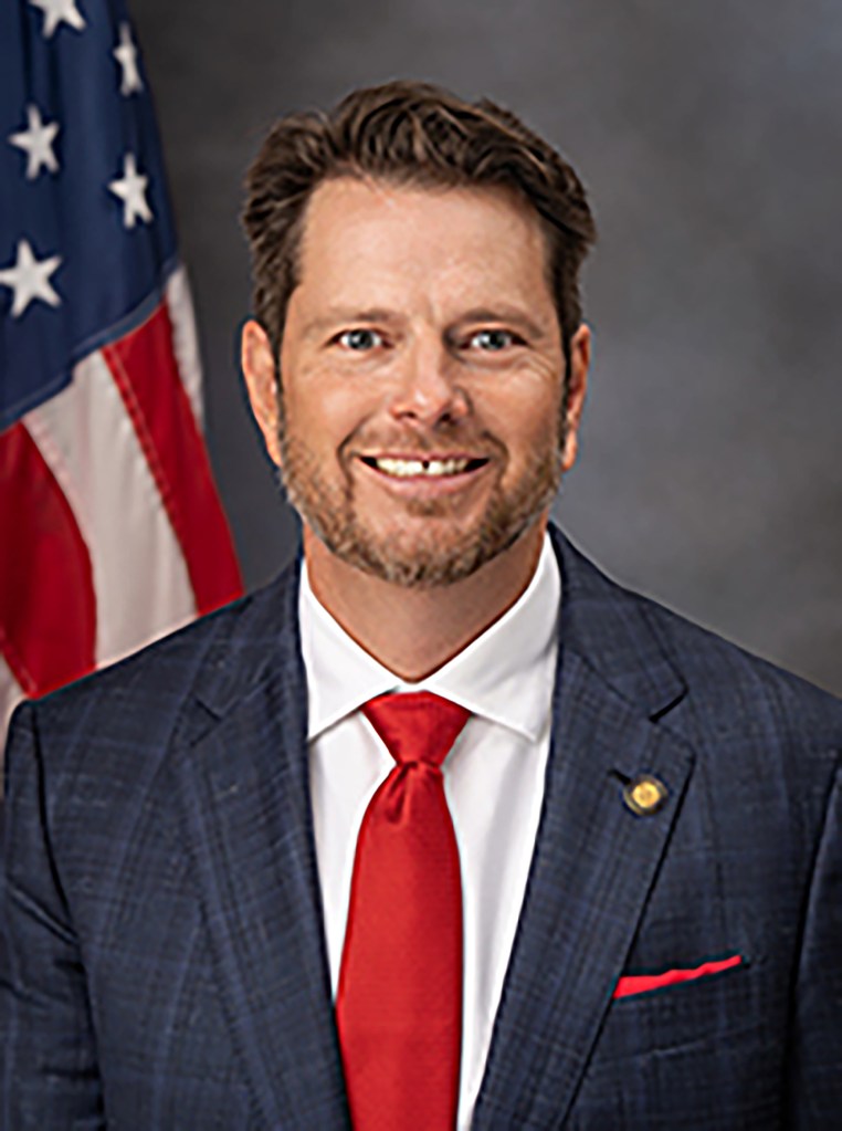 Headshot of a man with brown hair and a beard, wearing a blue plaid suit, white shirt, and red tie, with an American flag in the background.