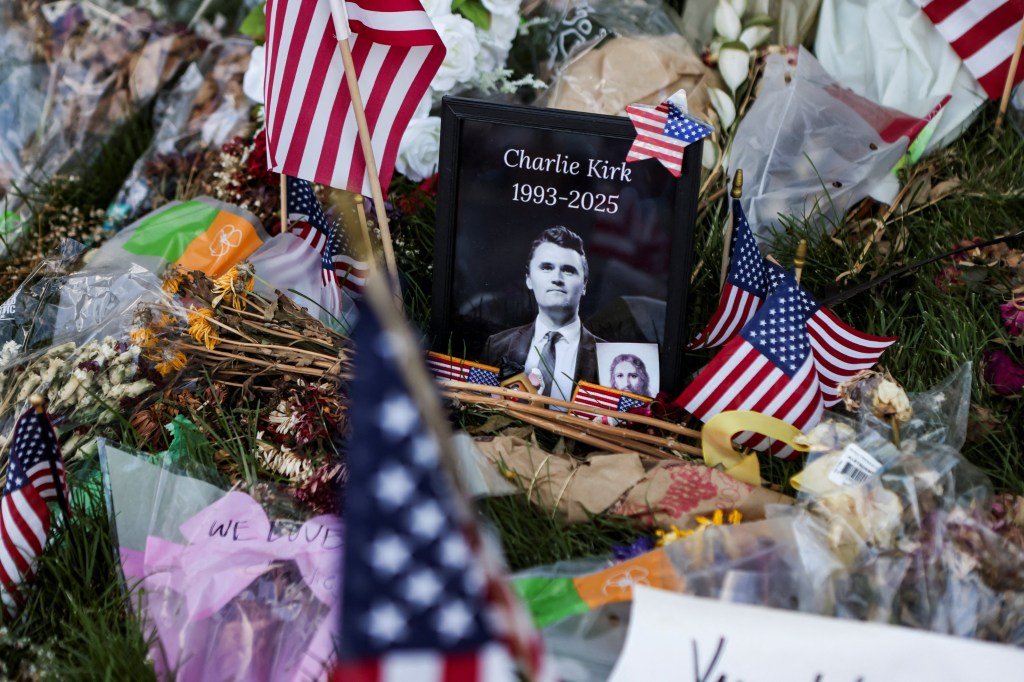 A memorial with flowers and American flags surrounding a framed photo of Charlie Kirk, with his birth and death years as 1993-2025.