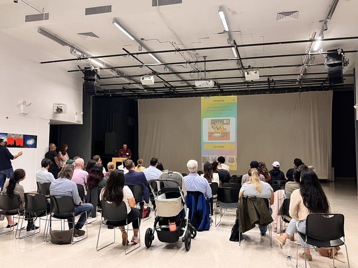 A group of people watch a presenter speaking at a podium with a colorful slide projected on a large screen in a bright, modern room.