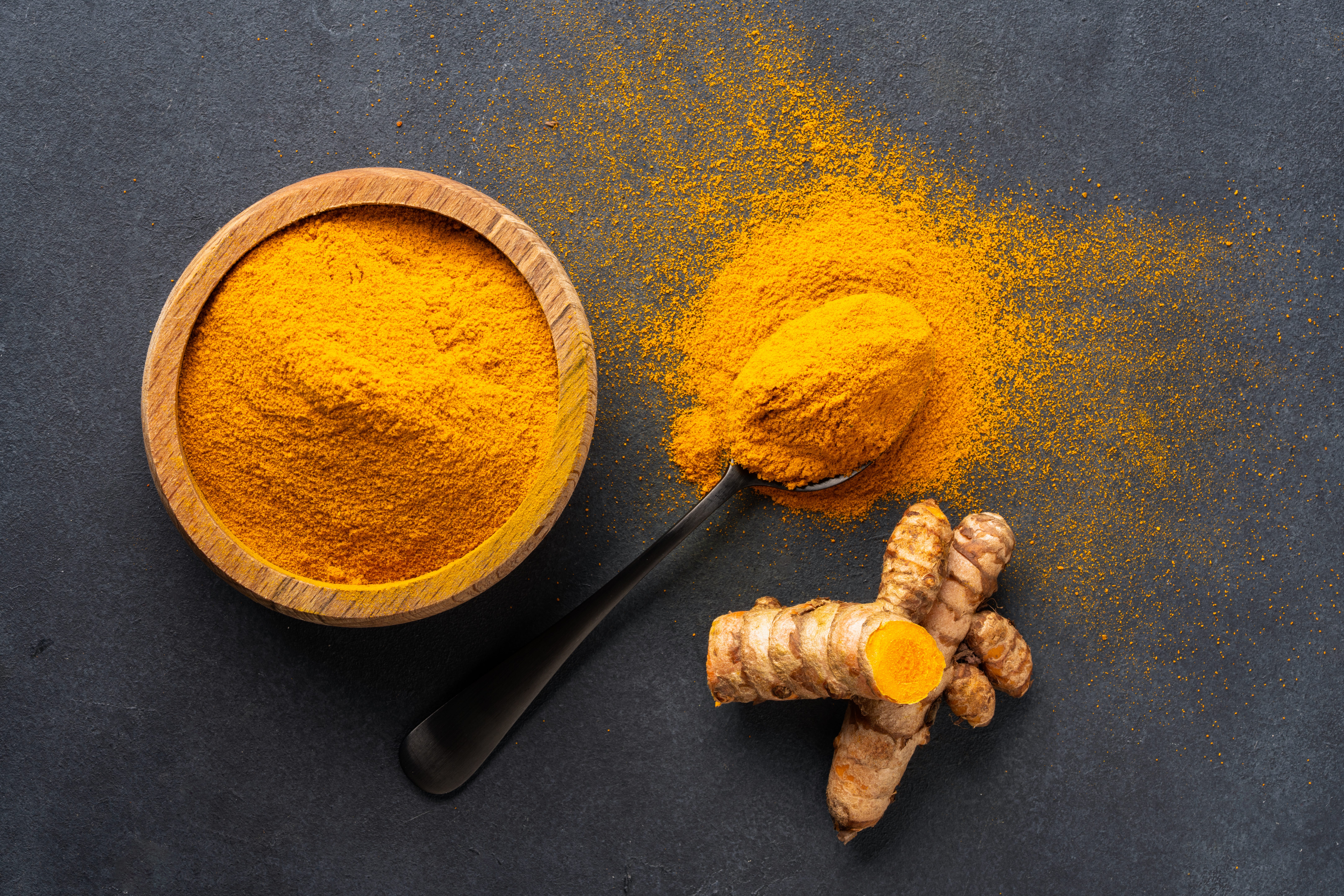 bright orange turmeric powder in a bowl and scattered across a black background next to a piece of cut turmeric root.