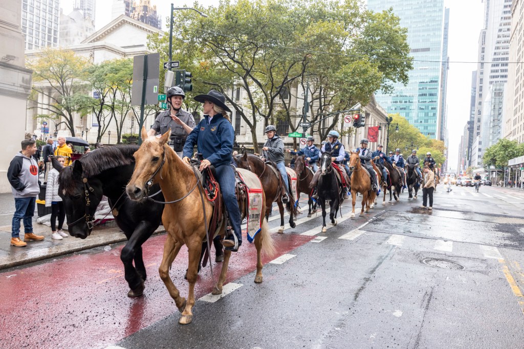 A line of people on horseback ride down a city street, with one woman in a cowboy hat speaking to a police officer.