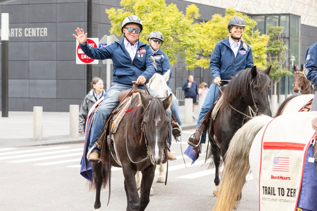 A line of people on horses in blue BraveHearts jackets ride down a street, with one rider waving to the left.
