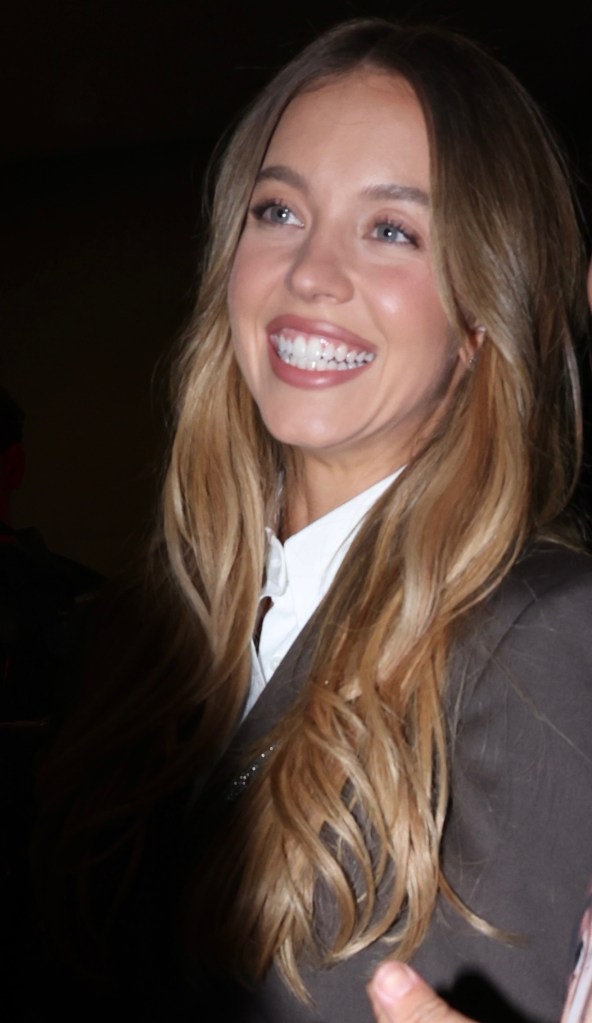 Sydney Sweeney smiling while wearing a brown blazer and white blouse.