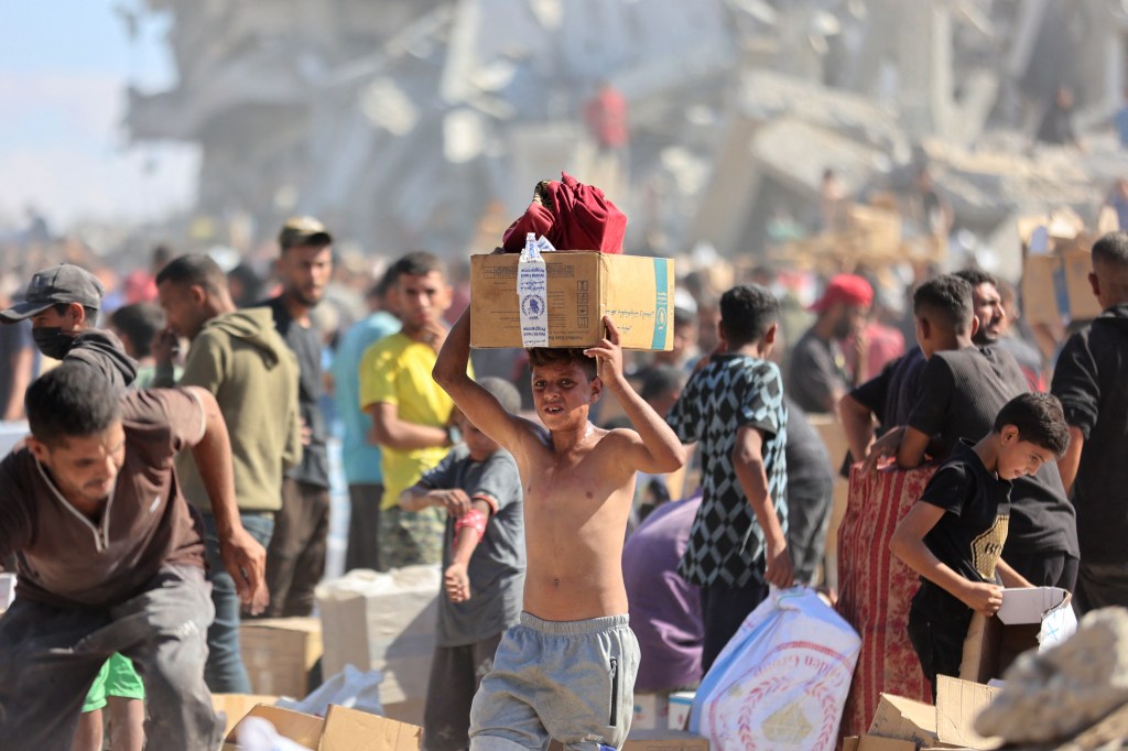 A young boy carries a box of aid from the World Food Programme on his head amidst a crowd of people in a war-torn area.