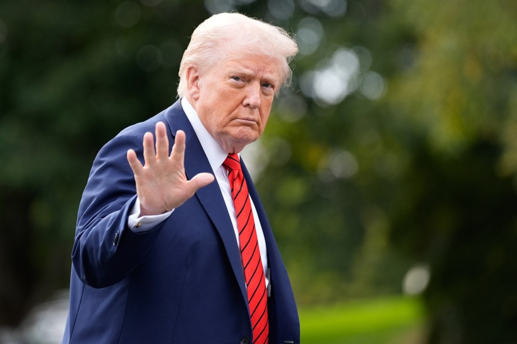 President Donald Trump walks on the South Lawn, hand raised, wearing a blue suit and a red and black striped tie.