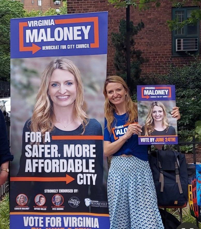 Virginia Maloney campaigning for City Council, holding a small sign and standing next to a large campaign sign.