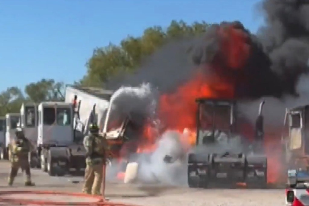 Two firefighters combat a truck fire, with thick black smoke rising above.