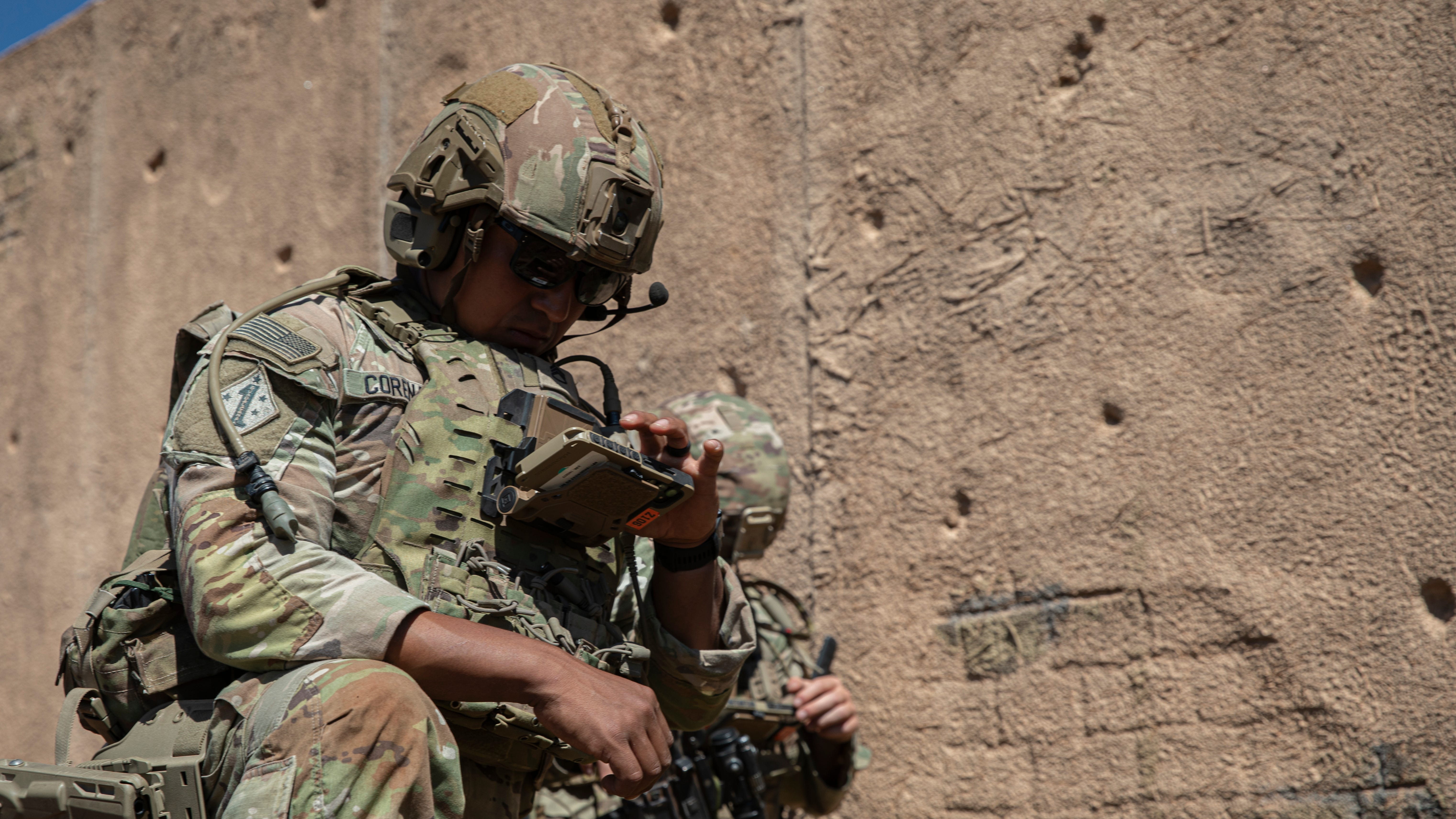 A soldier wearing camouflage holds a small device attached to his chest, looking down at the device. In the background is brown wall of clay and brick.