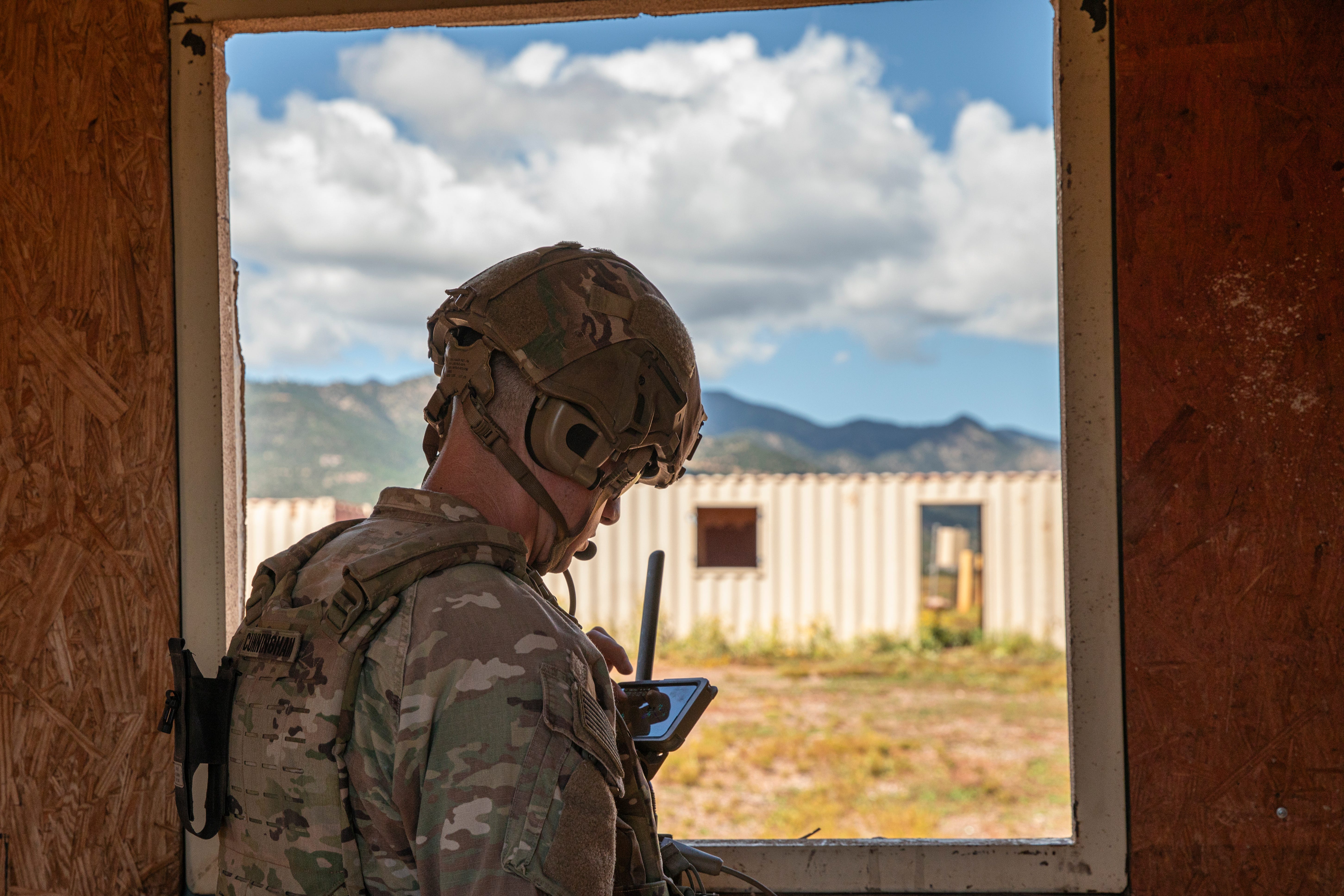 A soldier wearing camouflage looks down at an electronic device. He is framed by a window looking out onto an Army field training area with mountains and a cloudy blue sky in the background.