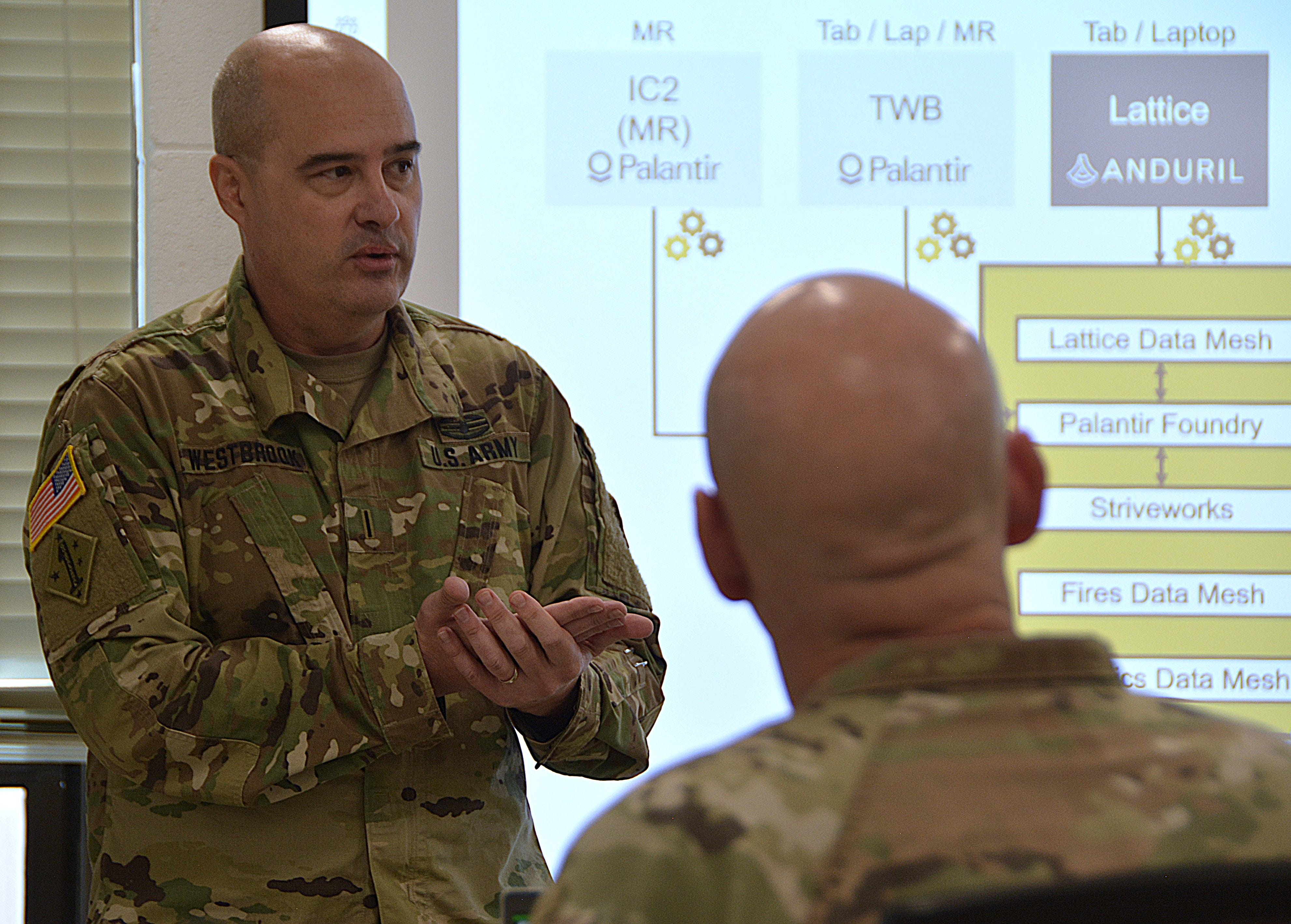 A man wearing camouflage stands in front of a projection screen that shows the breakdown of the US Army's new command and control system. In the foreground is the back of a man's head.