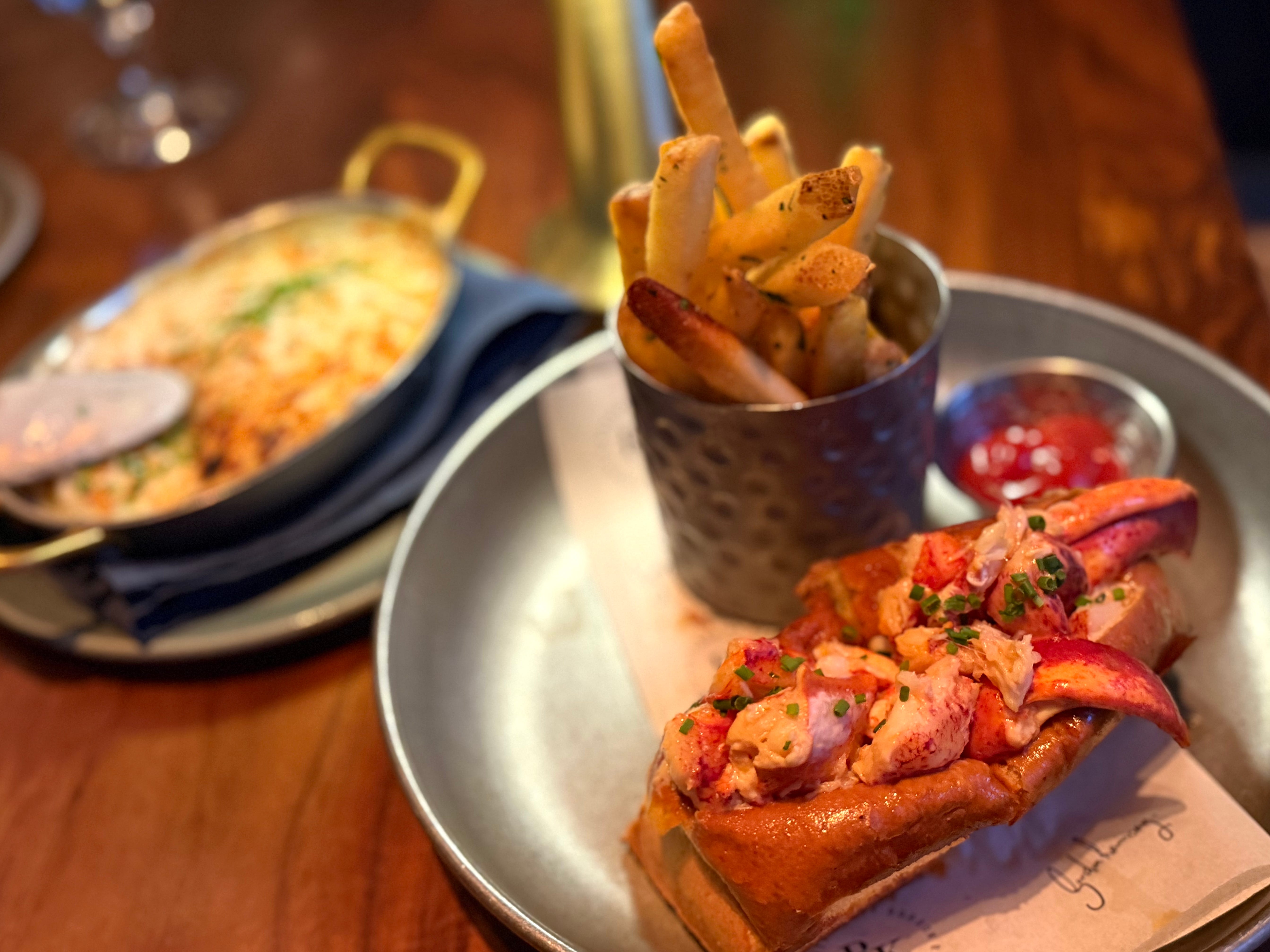 A lobster roll on a plate with a cup of fries and a potato gratin side dish in the background.