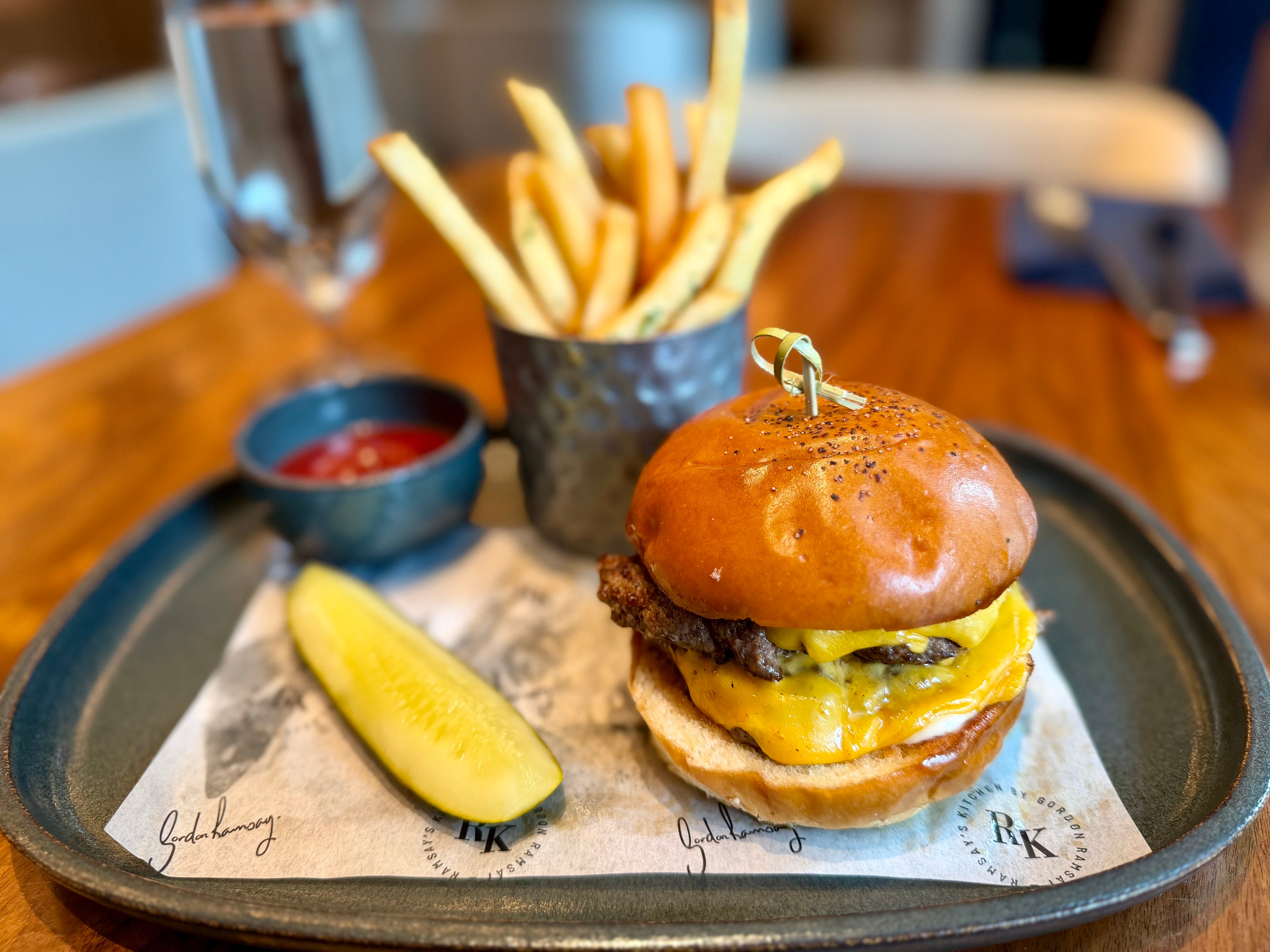 A plate with a burger, a cup of fries, and a side of ketchup, and a pickle on a plate at Ramsay's Kitchen.
