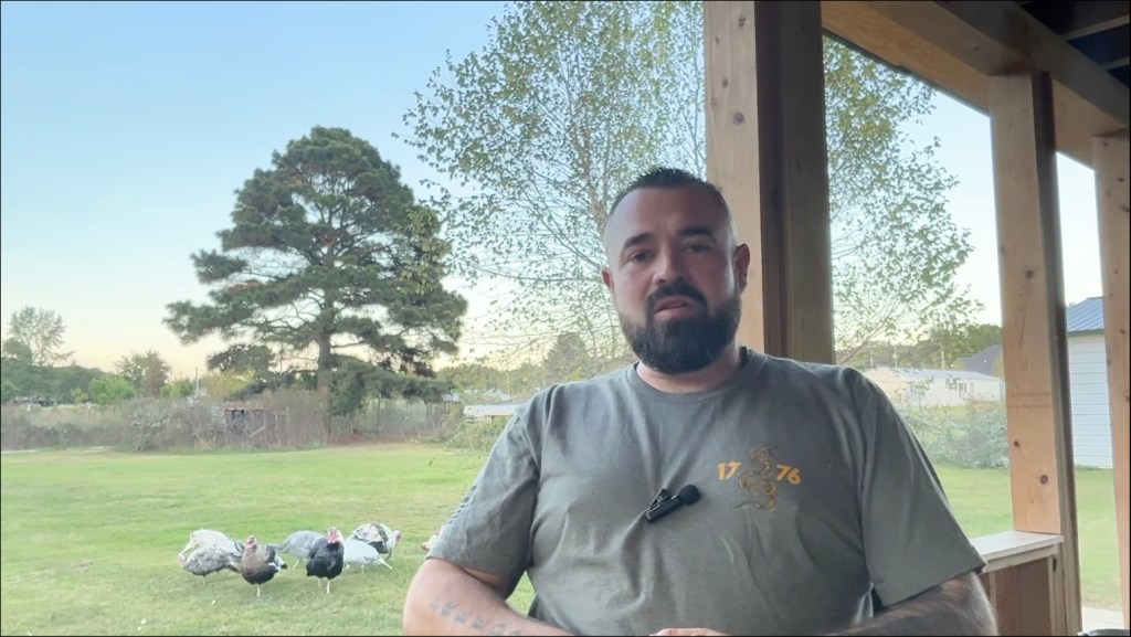 A man with a beard stands on a porch with turkeys in the background.