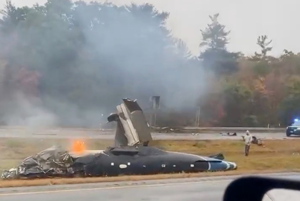 A crashed small plane smoking on the side of a highway.