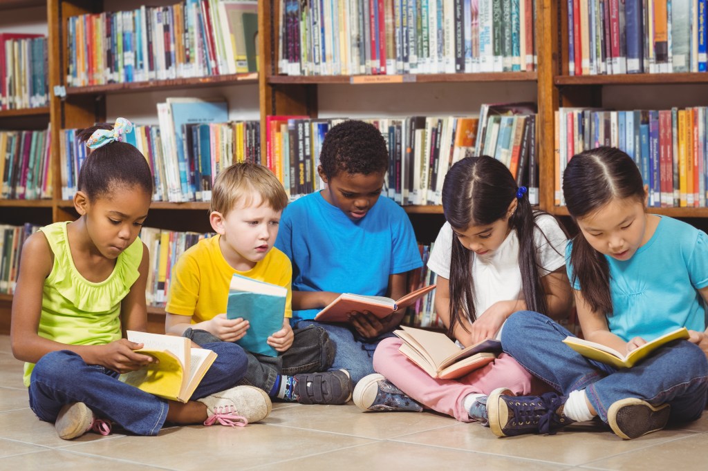 Five diverse elementary school pupils sitting on the library floor reading books.
