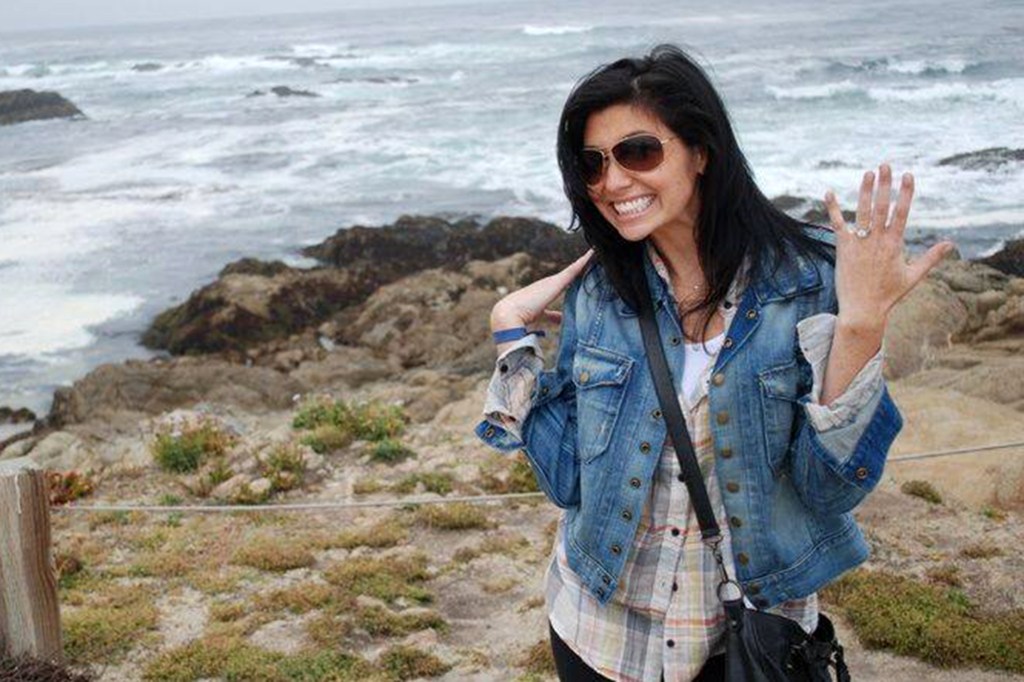 Ellen Greenberg smiling and showing an engagement ring, standing in front of an ocean with rocks.