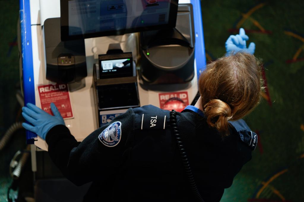A TSA worker in blue gloves screens a traveler at a security checkpoint.