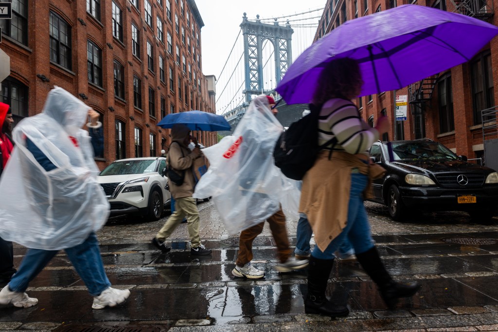 People walking with umbrellas and ponchos in the rain in Brooklyn with the Manhattan Bridge in the background.