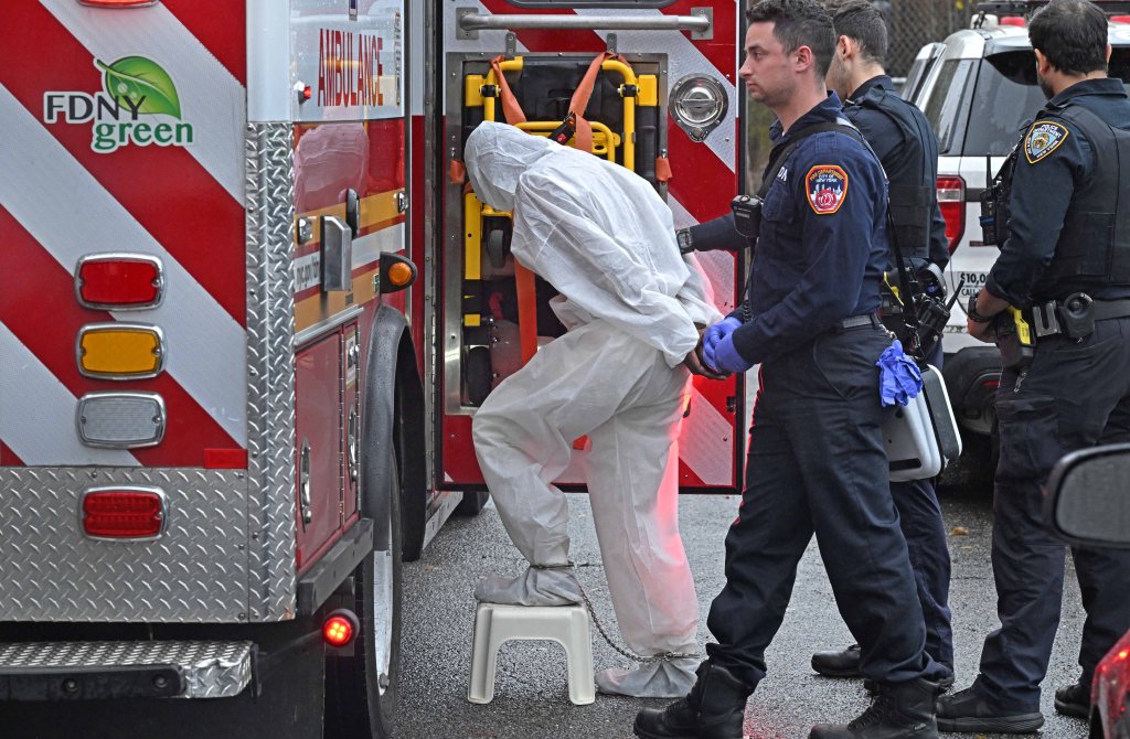 A person in a white protective suit and ankle shackles steps onto a stool while being escorted by a New York City Fire Department paramedic toward an ambulance, with a New York City Police Department officer nearby.