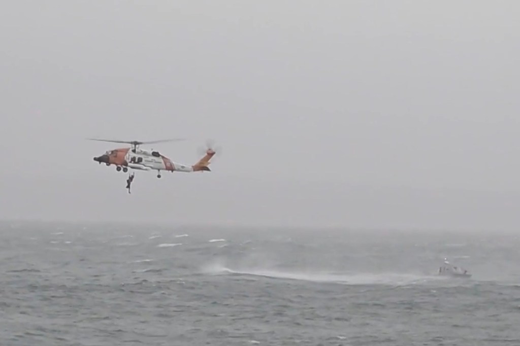A helicopter rescues a person from the ocean as a small boat nearby battles choppy waters.