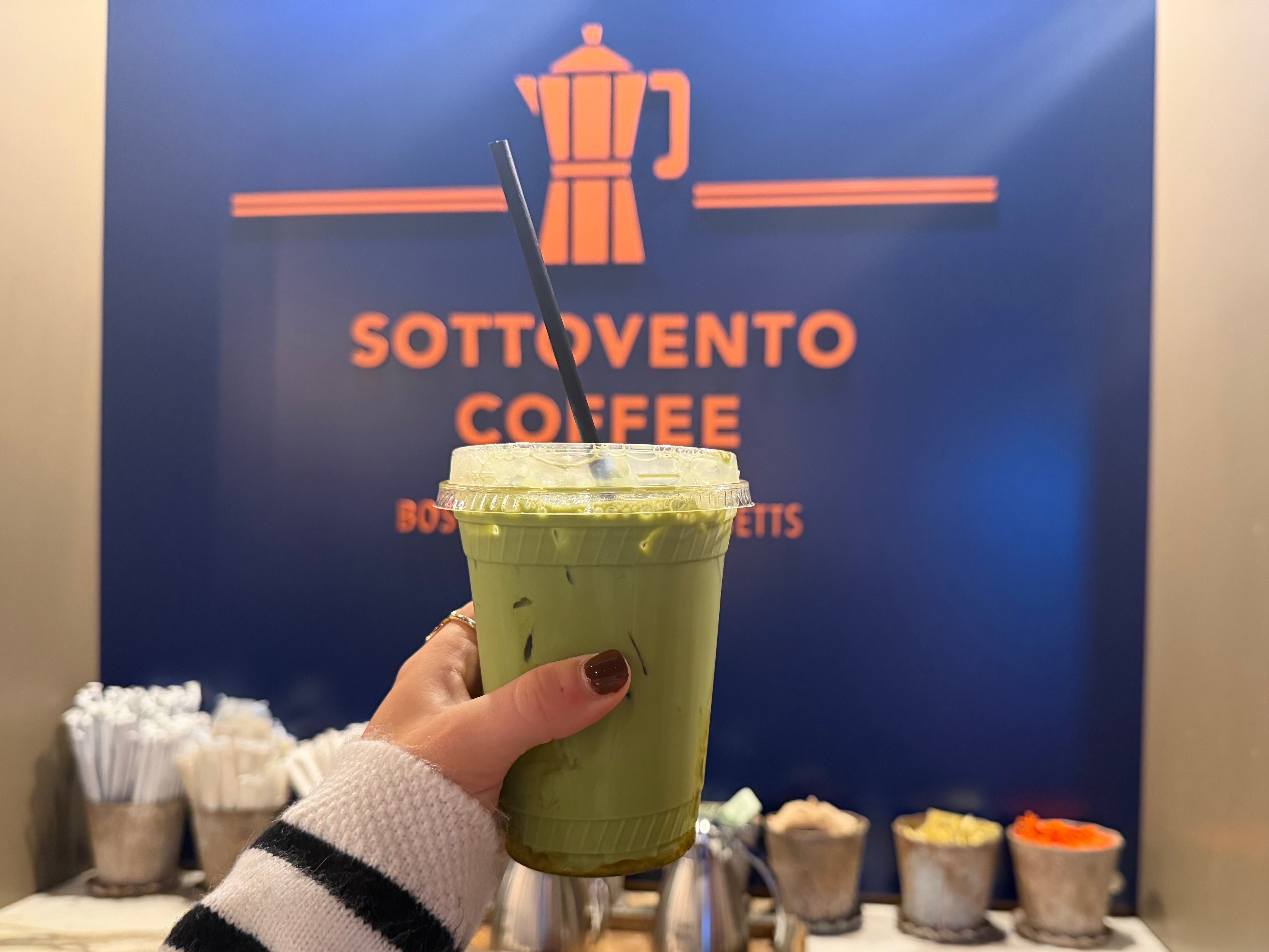 A hand holding up matcha in front of a Sottovento coffee sign.