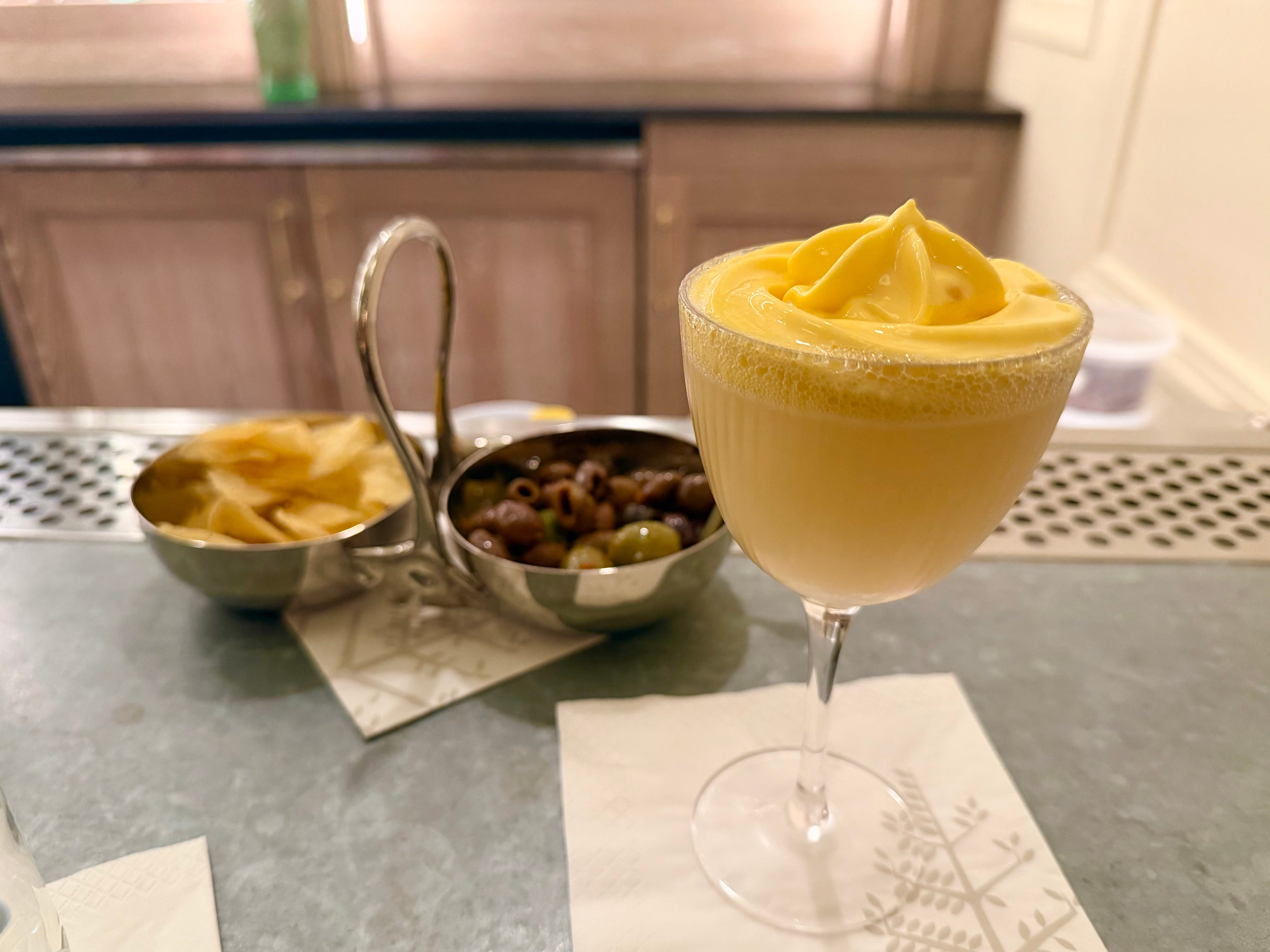 A yellow cocktail at a bar, with chips and olives in the background.