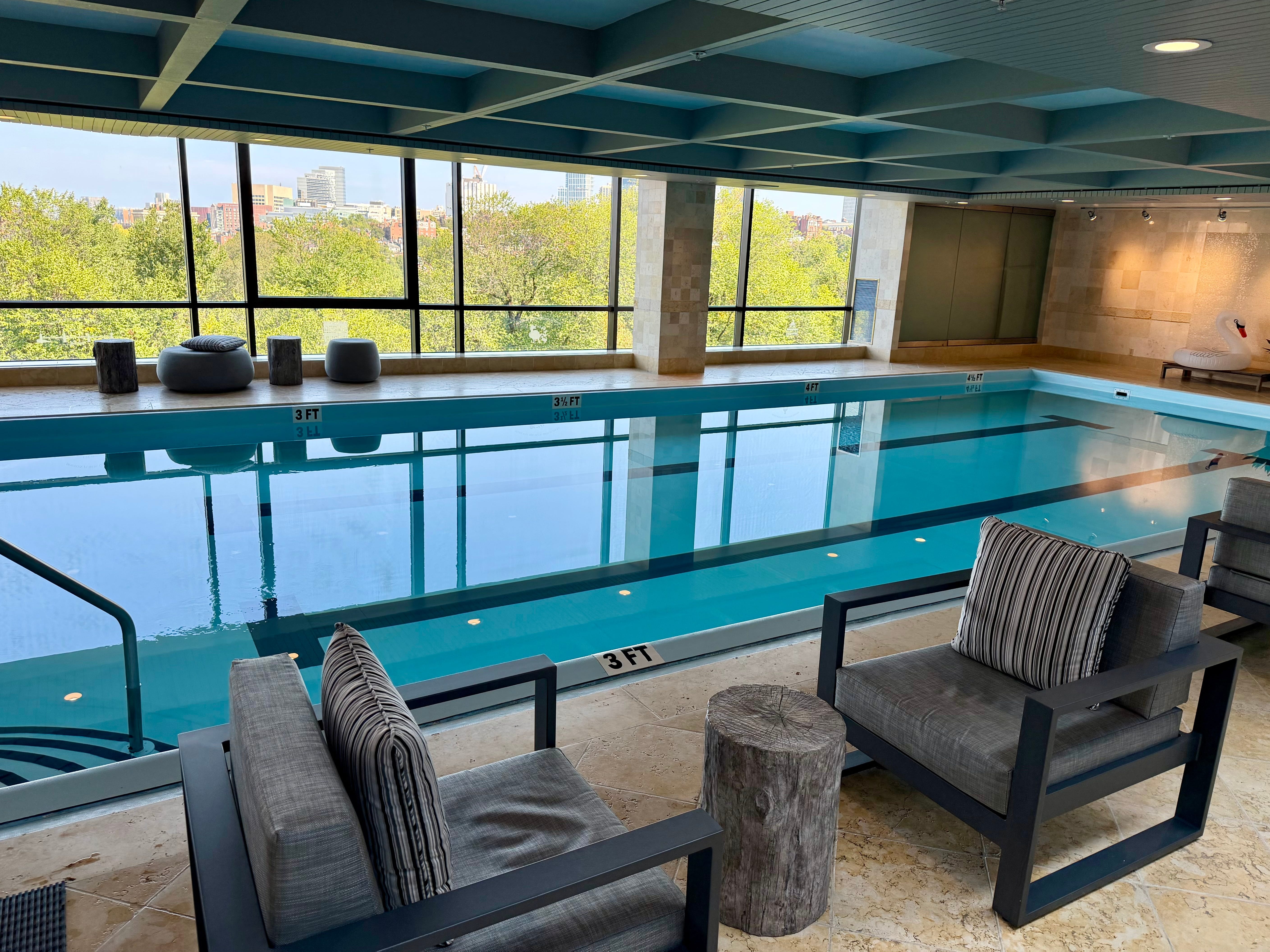 A blue pool at the Four Seasons, with nearby cushioned chairs and a view overlooking Boston.