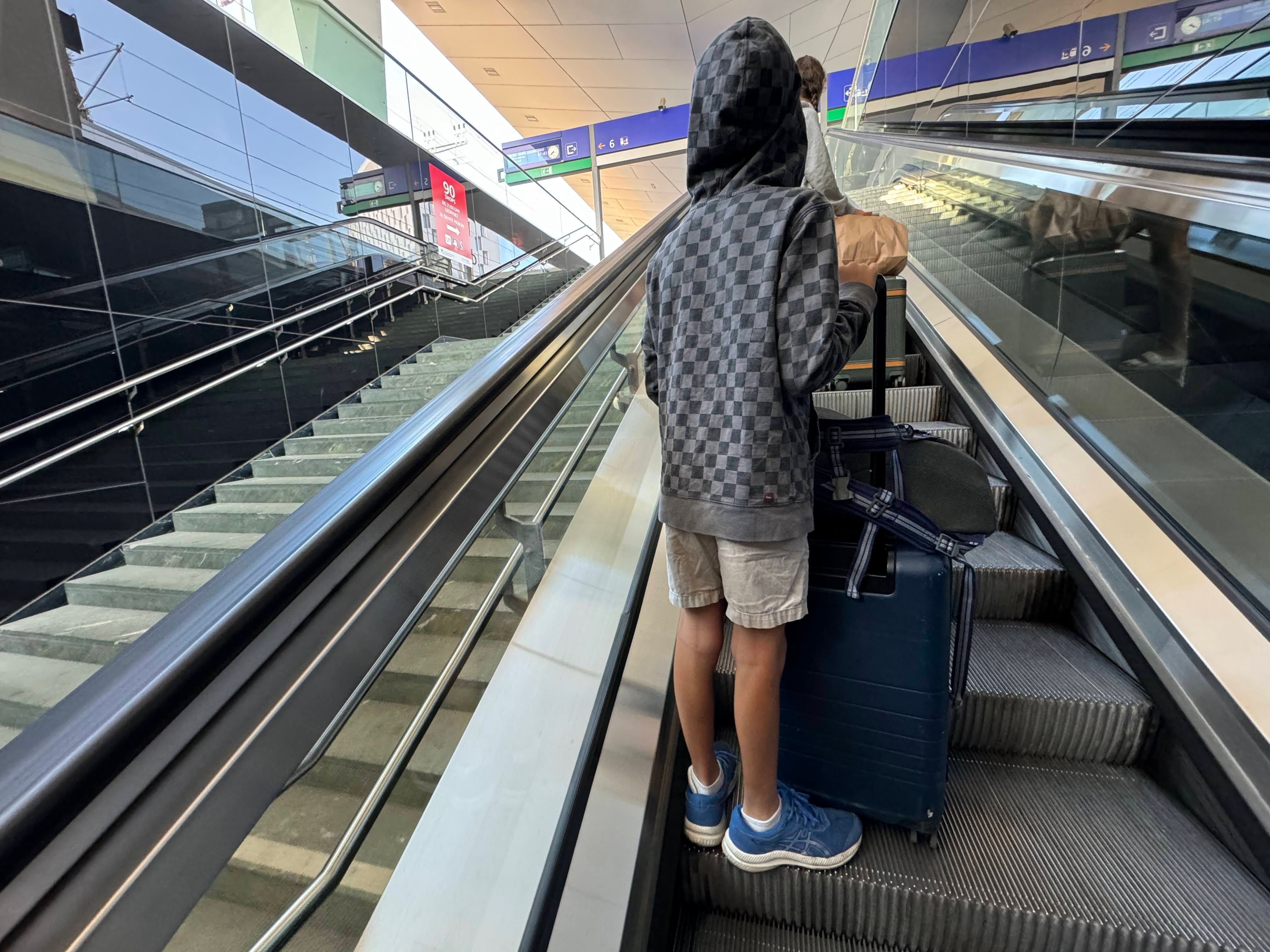 A child holding a suitcase and duffle bag on an escalator in a train station.