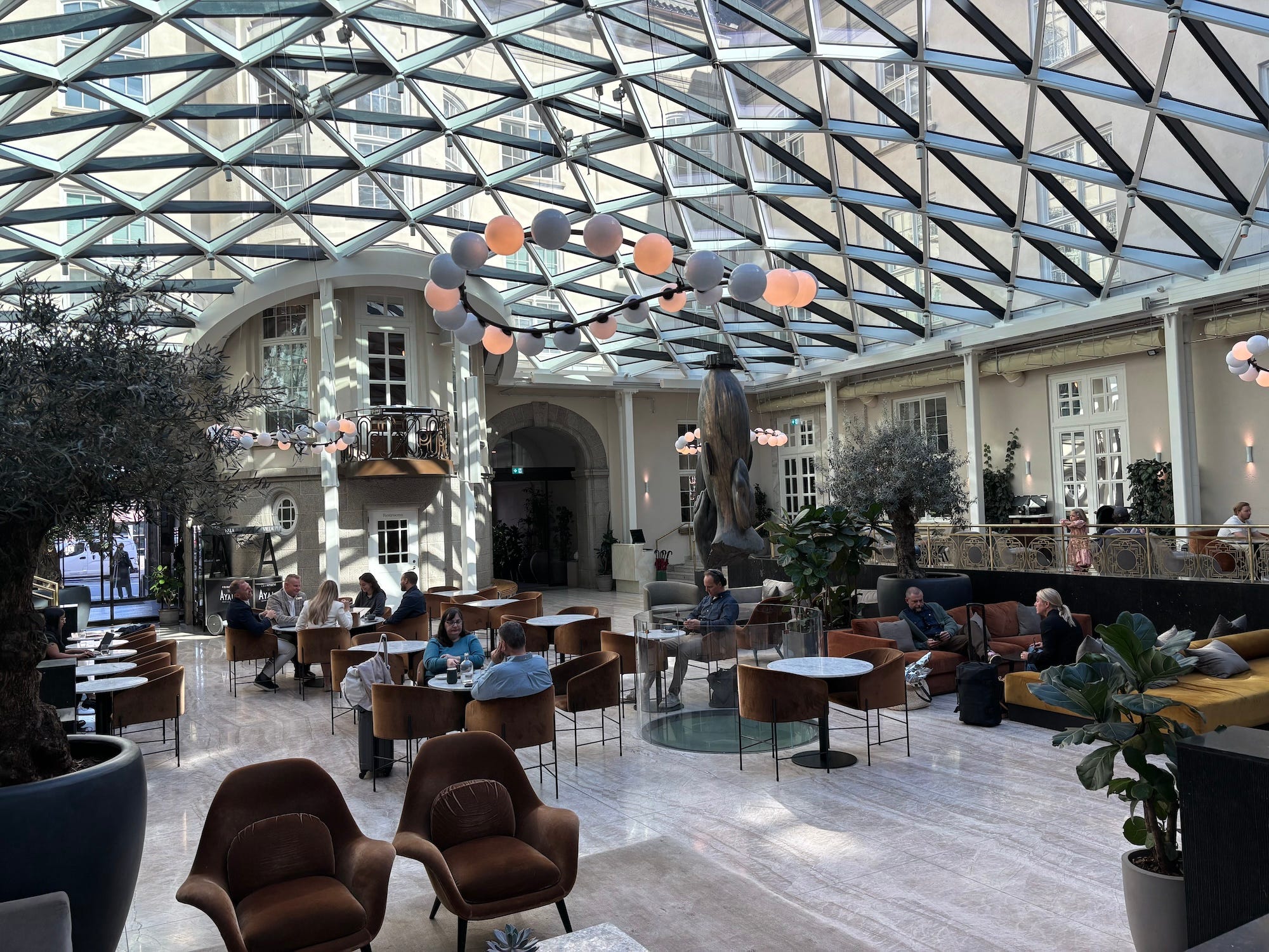 A lobby of a hotel with glass ceilings and an array of tables with guests sitting at them.