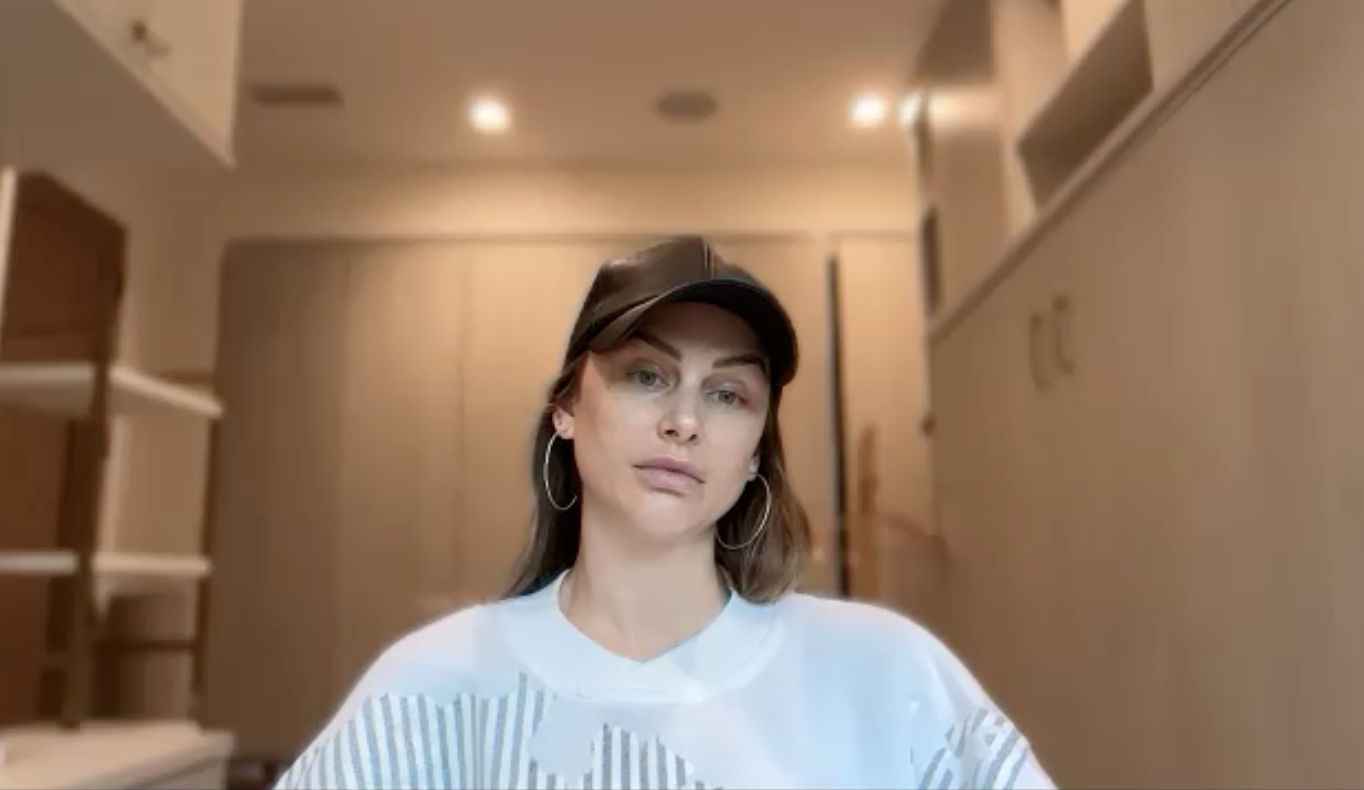 Lala Kent wearing a black hat, white shirt, and hoop earrings, looking at the camera.