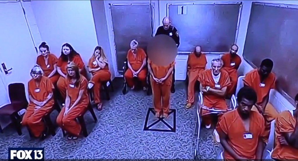 Inmates in orange jumpsuits sitting and standing in a room, with one inmate being escorted by a guard.