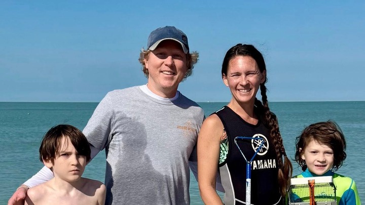 A family of four, two adults and two children, smiling at the camera with the ocean in the background.