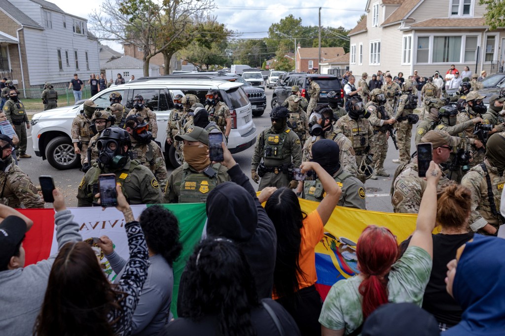 Federal agents in tactical gear and gas masks face off against community members holding up flags, some filming with phones.