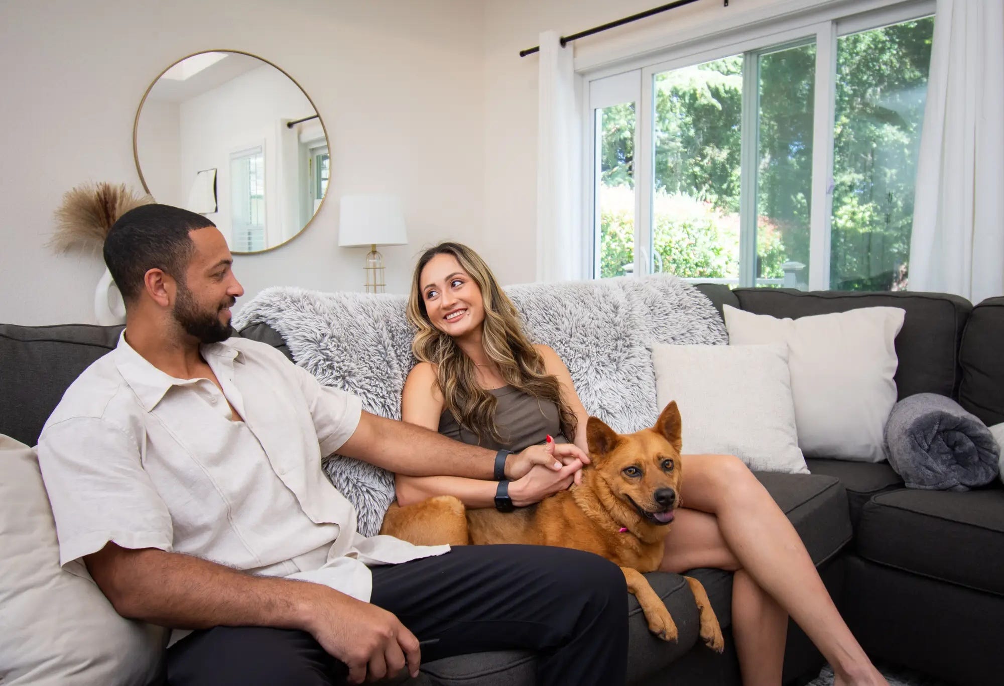 Aislyn and Ali Benjamin are inside their house, sitting on a couch with their dog.