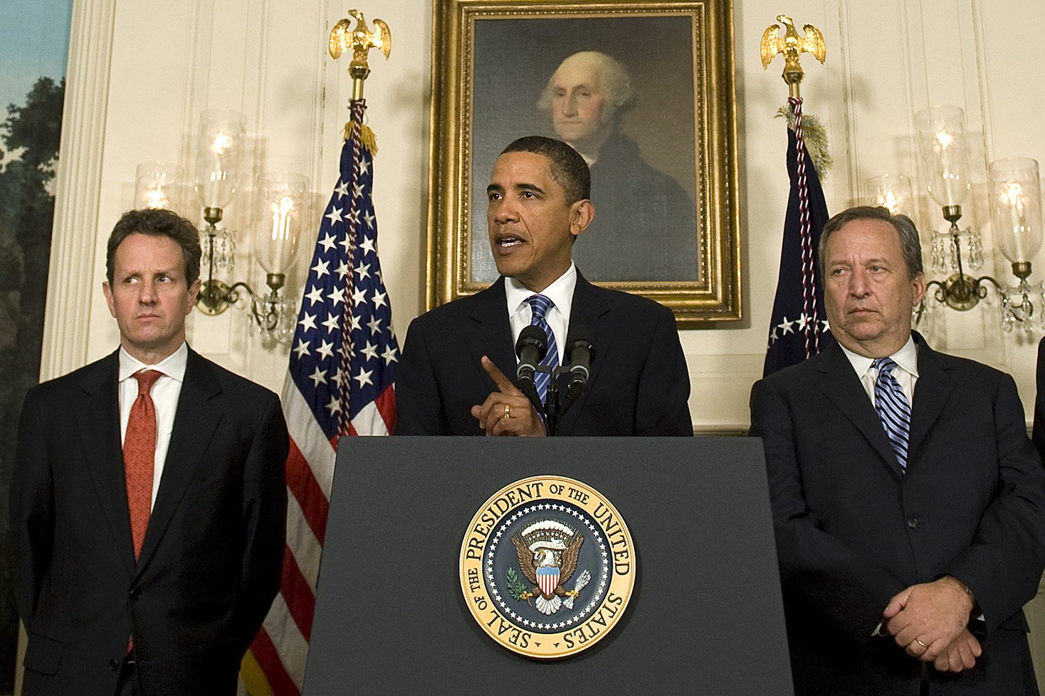 Barack Obama, dressed in a black suit and tie while standing at a podium affixed with the presidential seal, speaks between two men standing on either side. Both of the other men wear dark suits and ties. A portrait of George Washington hangs on the wall behind them.