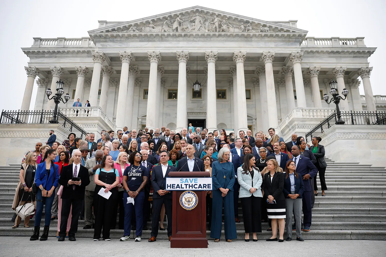 A group of dozens of U.S. lawmakers and supporters stand together on the steps of the Capitol, gathered behind a podium with a sign reading “Save Healthcare,