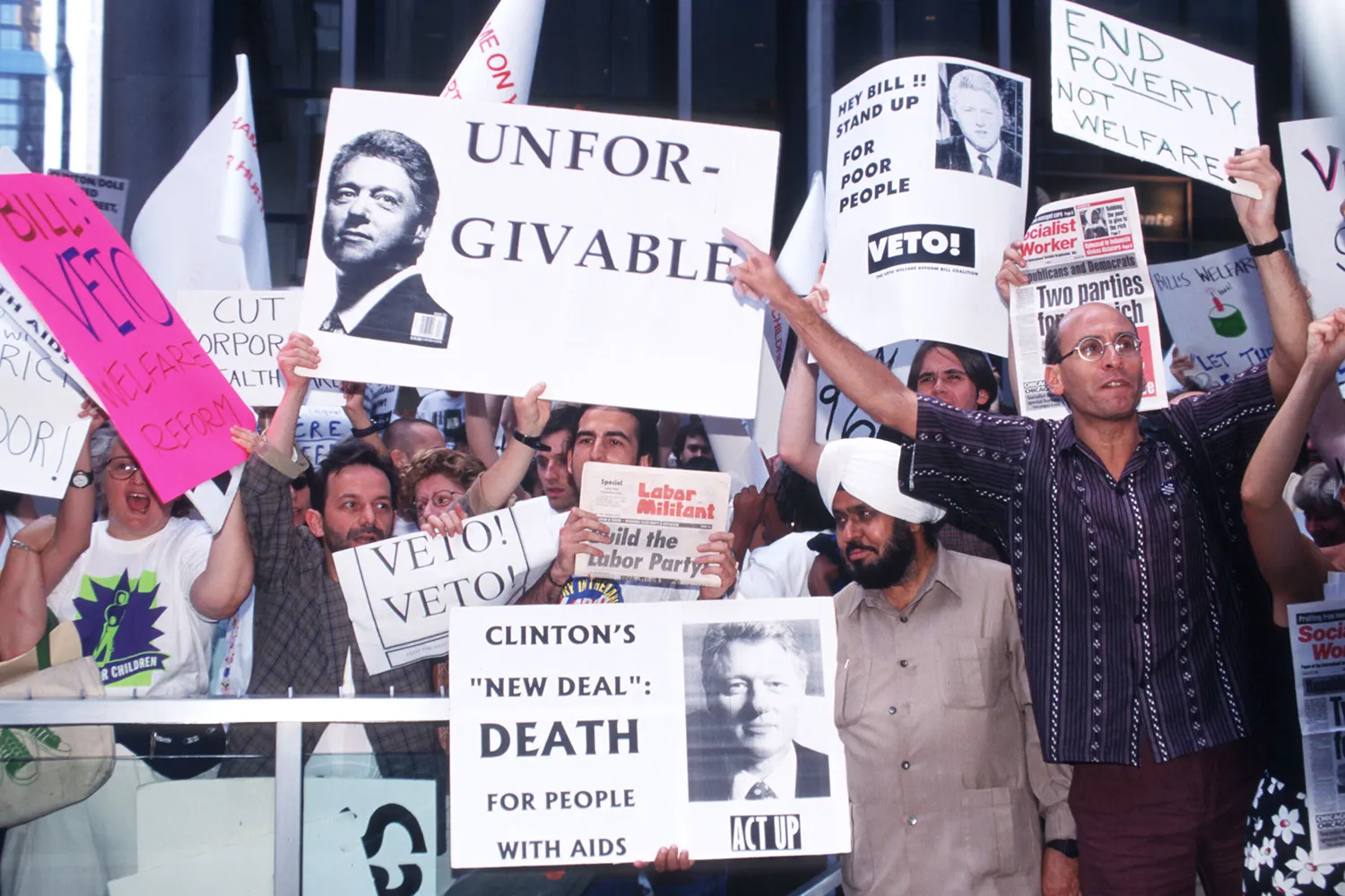 Protesters crowd against a barrier and hold signs criticizing Bill Clinton’s welfare reform, with messages such as “UNFORGIVABLE,” “VETO!” and “End Poverty Not Welfare,” during a demonstration.