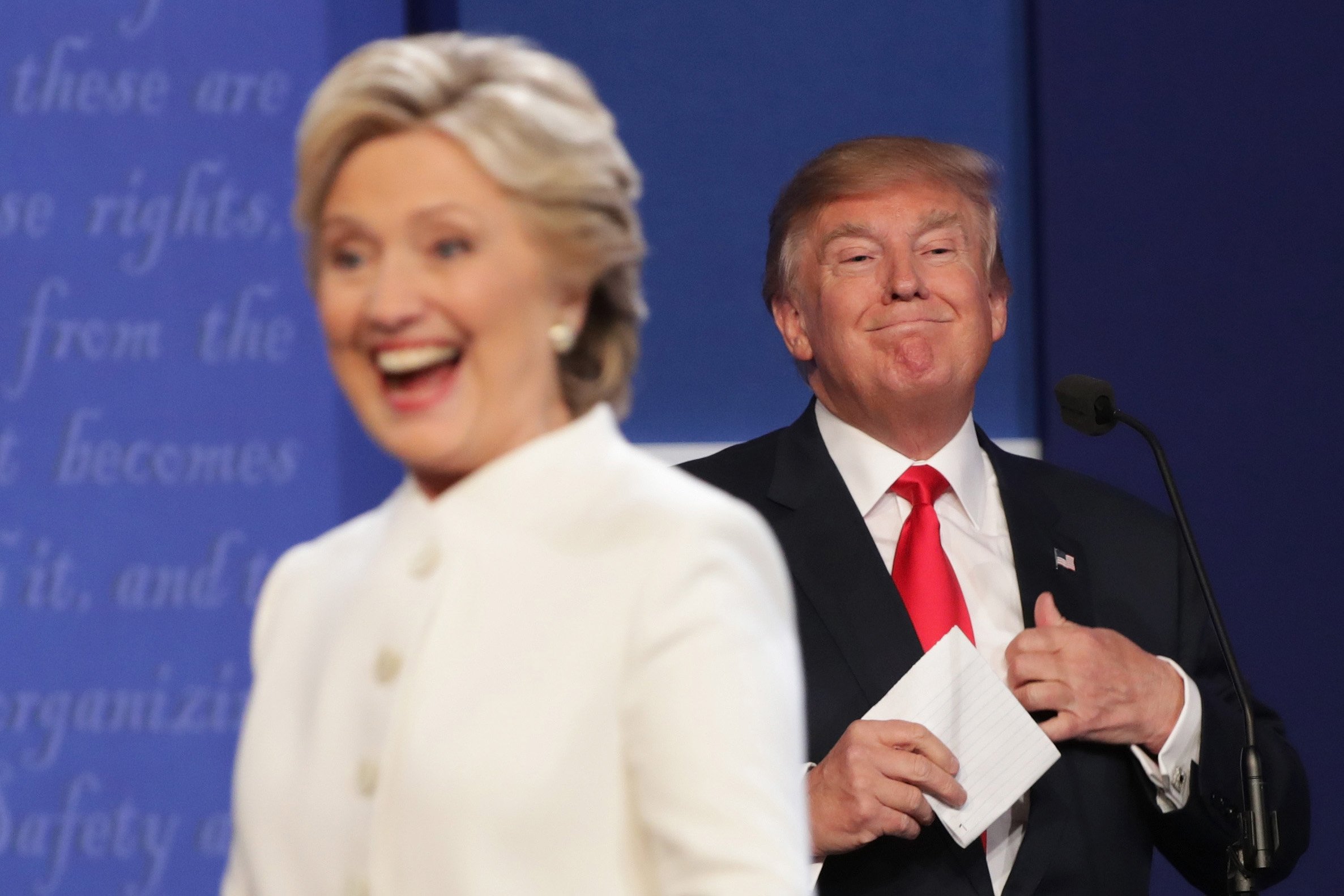 Hillary Clinton is seen out-of-focus in the foreground as she smiles while looking out of frame, while Donald Trump smiles behind her, in focus and looking the other way.