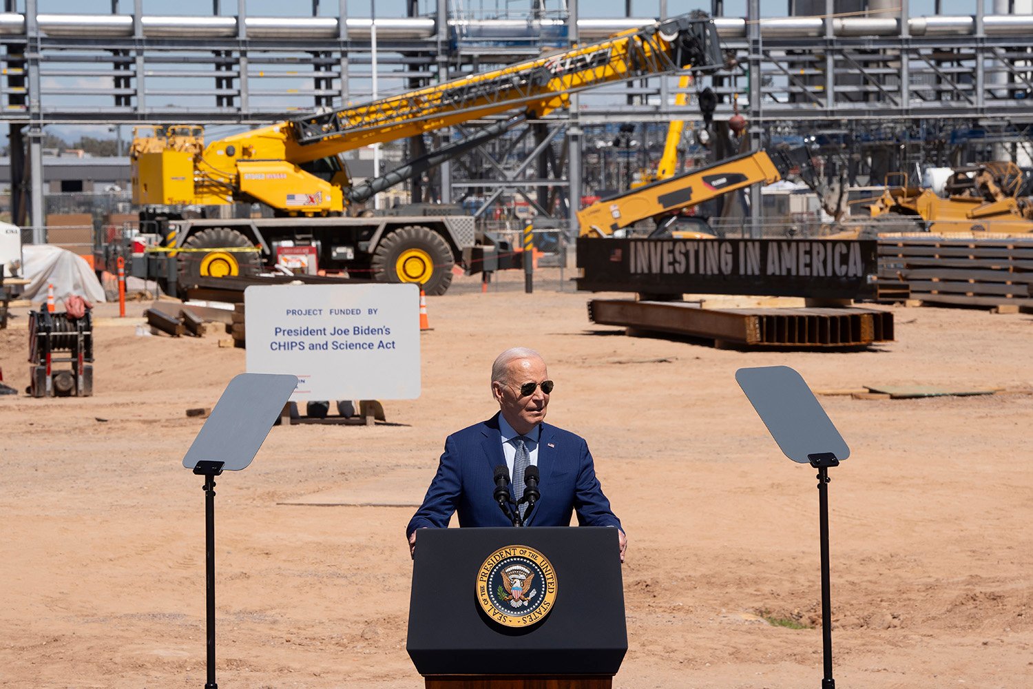 Joe Biden speaks at a podium in front of a large packed-dirt clearing clustered with construction machinery a plant in the desert. Biden wears a navy blue suit and sunglasses.