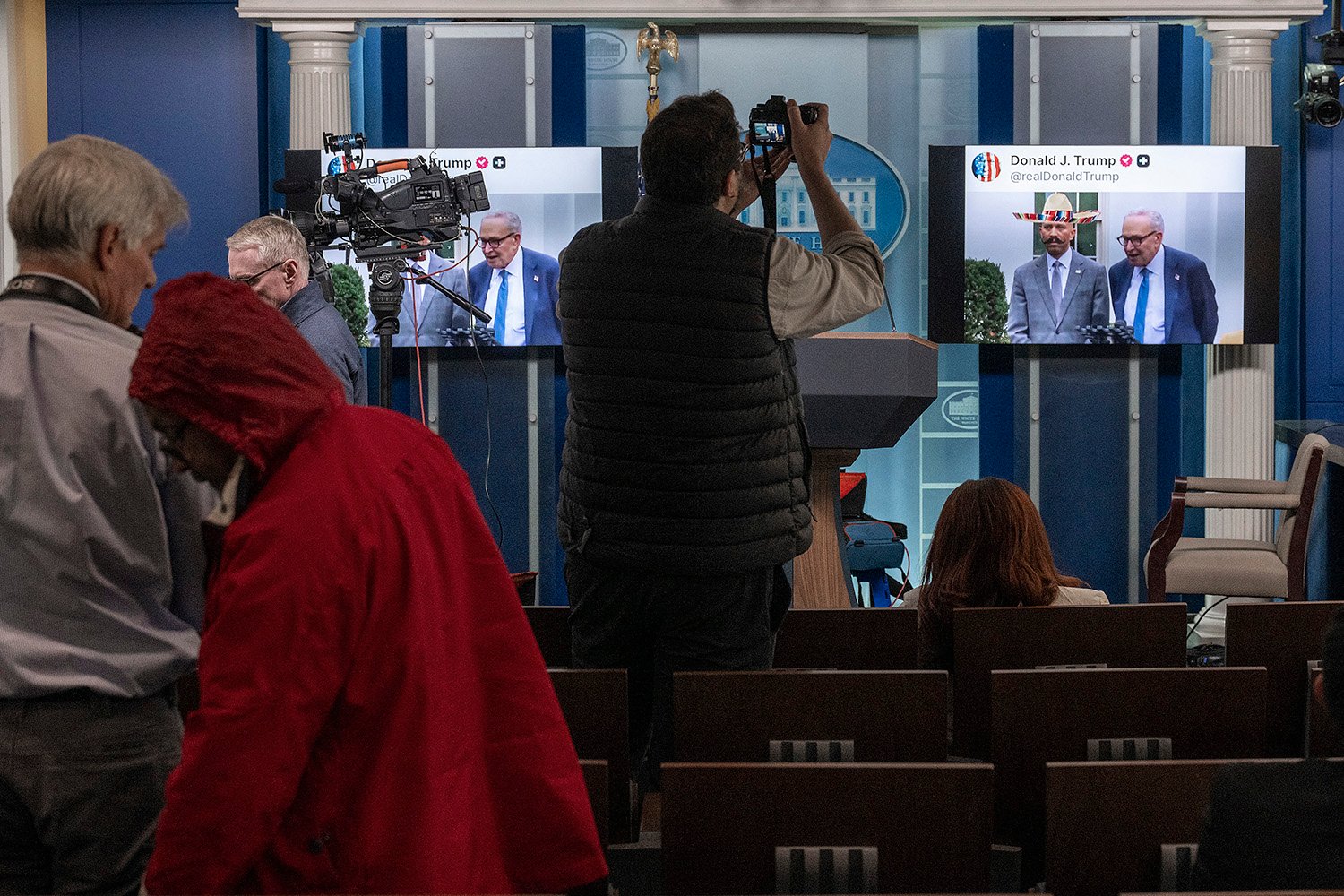 Journalists and photographers mill about in a briefing room lined with chairs, while on the far wall, two TV monitors play AI-generated videos mocking Democratic lawmakers.