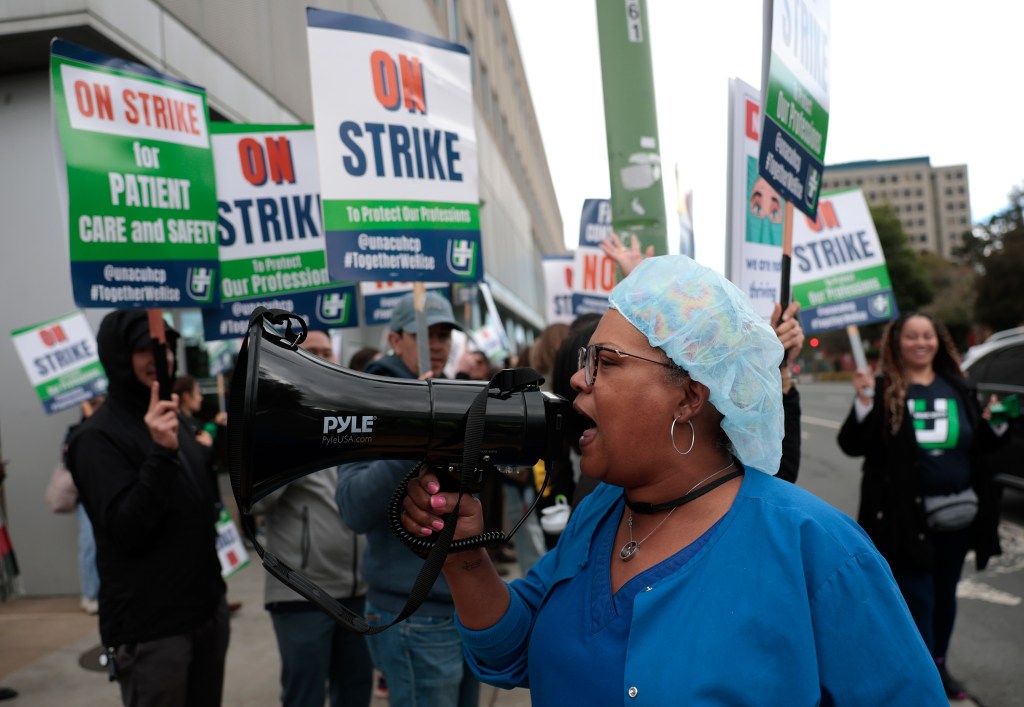 A Kaiser Permanente healthcare worker wearing a blue scrub and a blue hair net uses a megaphone during a strike.